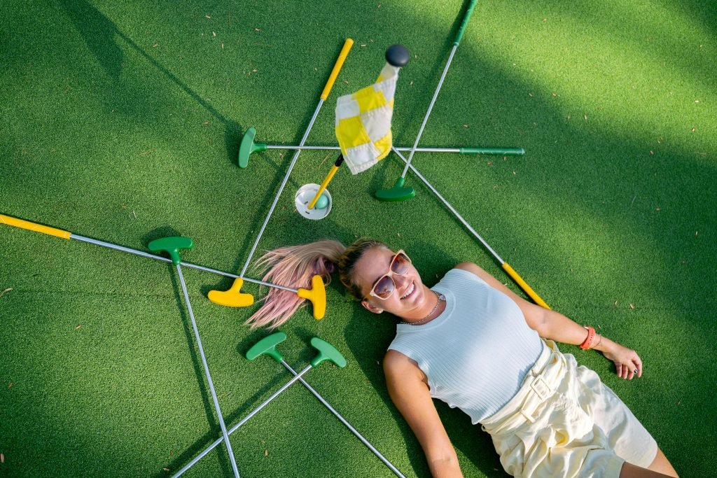 Woman enjoying a sunny day at the mini golf course, surrounded by putters and a flag.